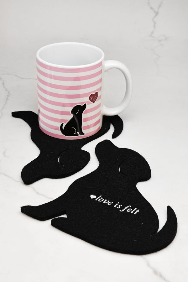 A white ceramic mug with pink horizontal stripes and a black puppy illustration sits on a black felt dog-shaped coaster. Another black felt dog-shaped coaster in the foreground has the words 'love is felt' in white script.
