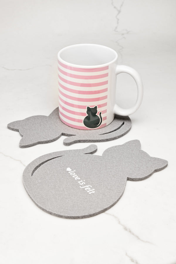 A white ceramic mug with pink horizontal stripes and a black cat illustration sits on a gray cat-shaped felt coaster, with two matching gray cat coasters on a light marble surface.