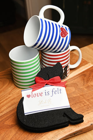 Three ceramic mugs with blue, green, and red stripes are stacked on a wooden surface next to a set of black felt dog coasters with a red ribbon and a card that reads 'love is felt' 