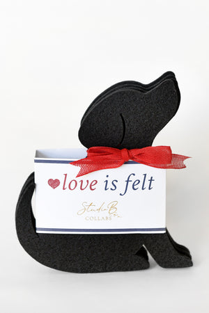 A black felt dog-shaped coaster set with a red ribbon bow, shown in front of a white gift box labeled 'love is felt' and 'Studio B. Collabs' on a clean white background.