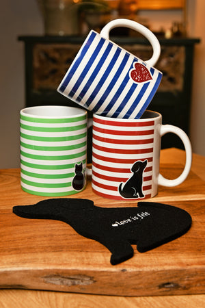 Three ceramic mugs with colorful horizontal stripes (green and white, red and white, blue and white) are stacked on a wooden table, with a black dog-shaped coaster in front featuring the text 'love is a felt'.