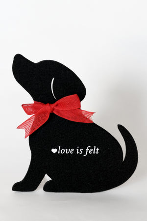 A black felt dog-shaped coaster with a red ribbon bow around its neck and the white text 'love is felt' on its side, set against a plain light gray background.