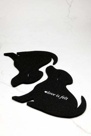 Two black felt dog coasters on a white marble background, with one dog featuring the white text 'love is felt' and a small heart.