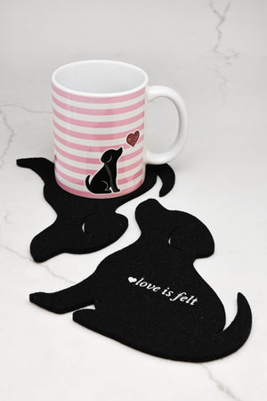 A white ceramic mug with pink horizontal stripes and a black puppy illustration sits on a black felt dog-shaped coaster. Another black felt dog-shaped coaster in the foreground has the words 'love is felt' in white script.