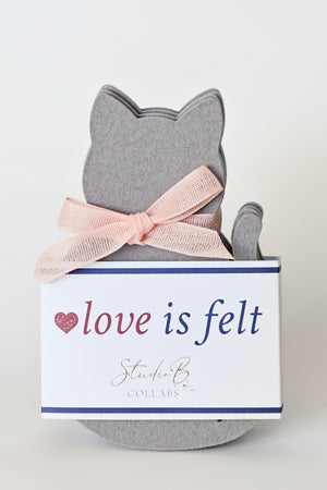 A gray felt cat-shaped decorative item tied with a pale pink ribbon and wrapped in a white gift box labeled 'love is felt' and 'Studio B Collars' on a clean white background.