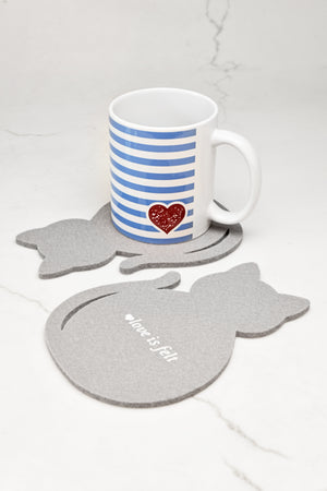 A white ceramic mug with blue horizontal stripes and a red glitter heart, resting on a gray cat-shaped felt coaster. Another matching gray cat coaster lies flat in the foreground on a white marble surface.