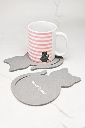 A white ceramic mug with pink horizontal stripes and a black cat illustration sits on a gray cat-shaped felt coaster, with two matching gray cat coasters on a light marble surface.
