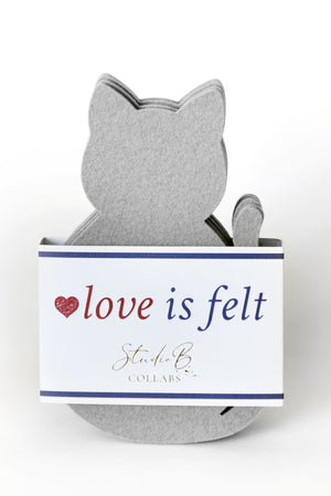 A gray felt cat-shaped decorative item displayed in front of a white branded card that reads 'love is felt' with Studio B Collabs branding.
