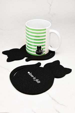 Oversized black cat coaster holding a white ceramic mug with green horizontal stripes and a black cat silhouette sits on a black cat-shaped coaster, with two additional black cat coasters in front on a white marble surface.