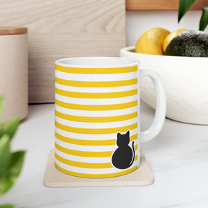 A white ceramic mug with yellow horizontal stripes and a black cat silhouette near the bottom, sitting on a beige coaster on a kitchen counter with a bowl of fruit in the background.
