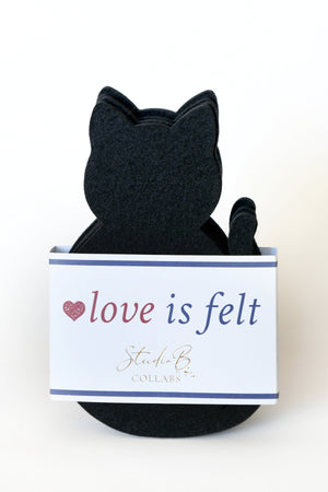 black cat coasters. these are felt cat-shaped coasters sitting inside a white gift box with the words 'love is felt’ printed on the front, set against a plain white background.