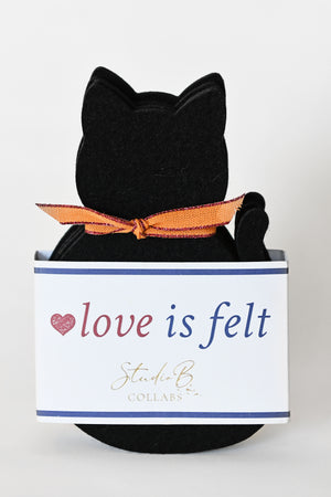 black cat coasters for Halloween. A black cat-shaped set of 4 coasters with a bright orange bow, displayed in front of a white branded box that reads 'love is felt’.