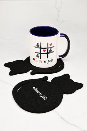 A white ceramic mug with a dark blue interior and handle sits on a black cat-shaped felt coaster, with two matching black cat coasters arranged on a white marble surface. The mug and coasters both feature the phrase "love is felt" with small cat and heart illustrations.