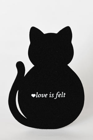 A black felt cat-shaped coaster viewed from the back, featuring the white printed text 'love is felt' with a small heart icon, set against a plain light gray background.