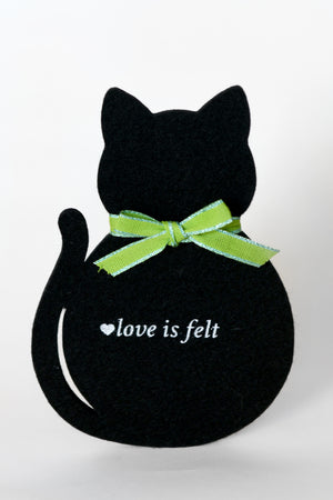 A black felt cat-shaped coaster with a bright green ribbon bow and the words 'love is felt' in white on the back, shown against a plain light background.